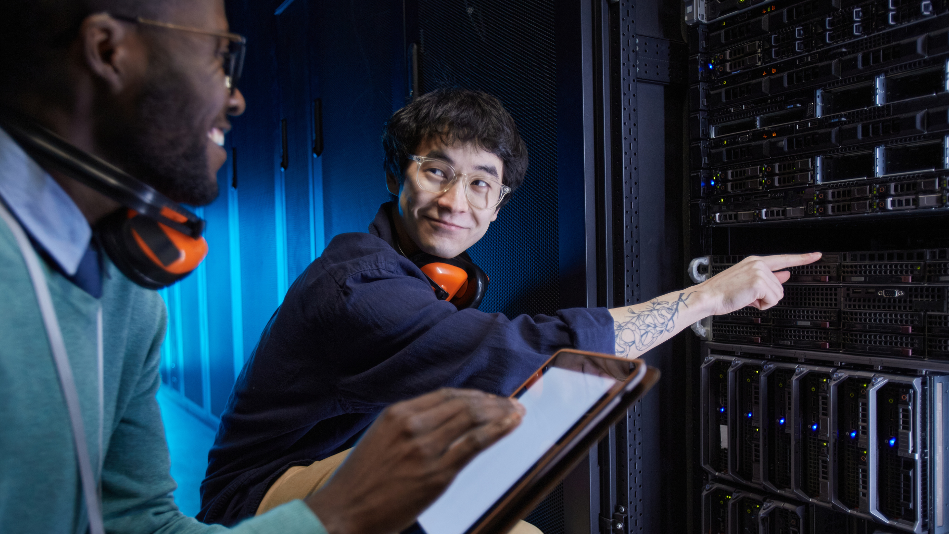 Two smiling data center technicians inspect server equipment. One person points to a server blade while the other holds a tablet, both wearing orange safety earmuffs around their necks.