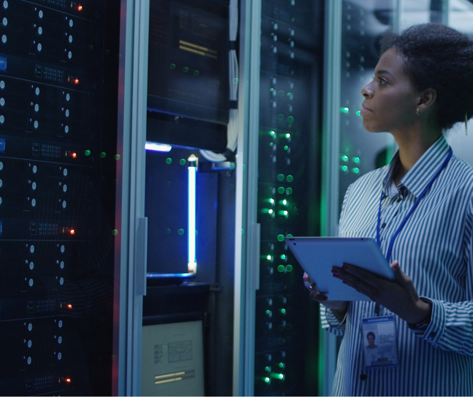 A woman wearing a striped shirt and ID badge stands in a dimly lit server room, holding a tablet and looking at a tall rack of networking equipment with red and green indicator lights.