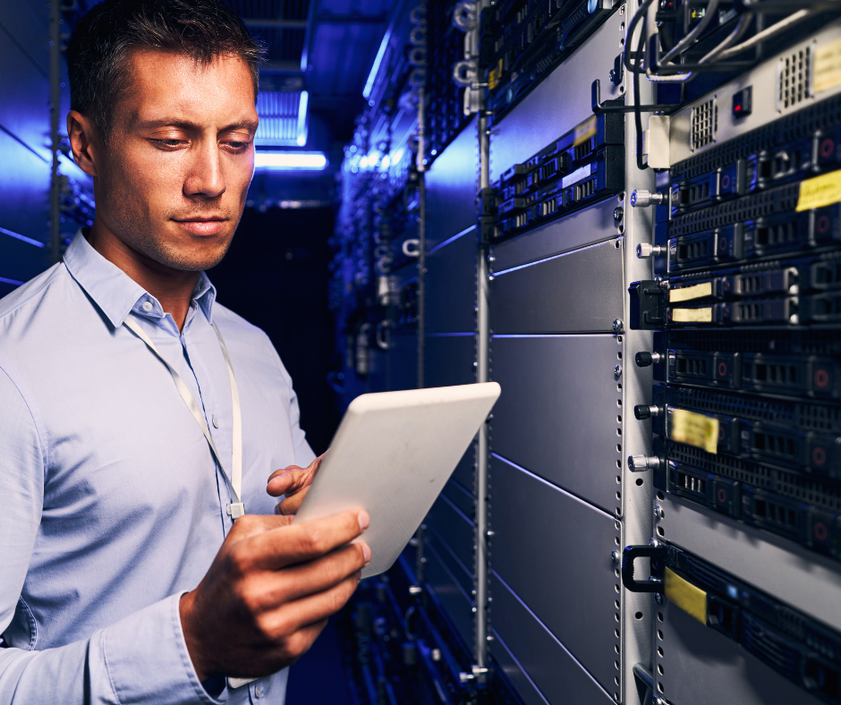 An IT professional is performing maintenance in a dimly lit data center, focusing on a white digital tablet while standing beside rows of servers and networking hardware. ups system