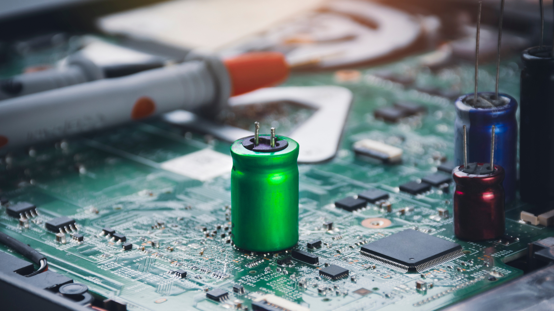 A close-up, detailed shot shows a bright green electrolytic UPS capacitors standing prominently on a complex green printed circuit board (PCB), surrounded by various smaller electronic components like integrated circuits (ICs) and surface-mount devices. To the right, smaller blue and red capacitors are visible, and the blurred background hints at technical work being performed, including a portion of a multimeter probe.